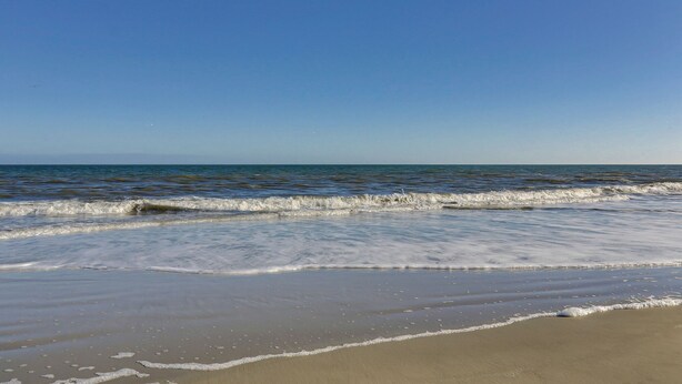 A beach with waves gently lapping onto the shore under a clear blue sky. - Sunset Rentals | Beachgate at Starfish 9 Dr. (Hilton Head Island)