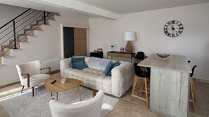 A modern living room with a sofa, chairs, a coffee table, a dining table with chairs, a clock on the wall, and a staircase. - Logement Maltus (Lussac)
