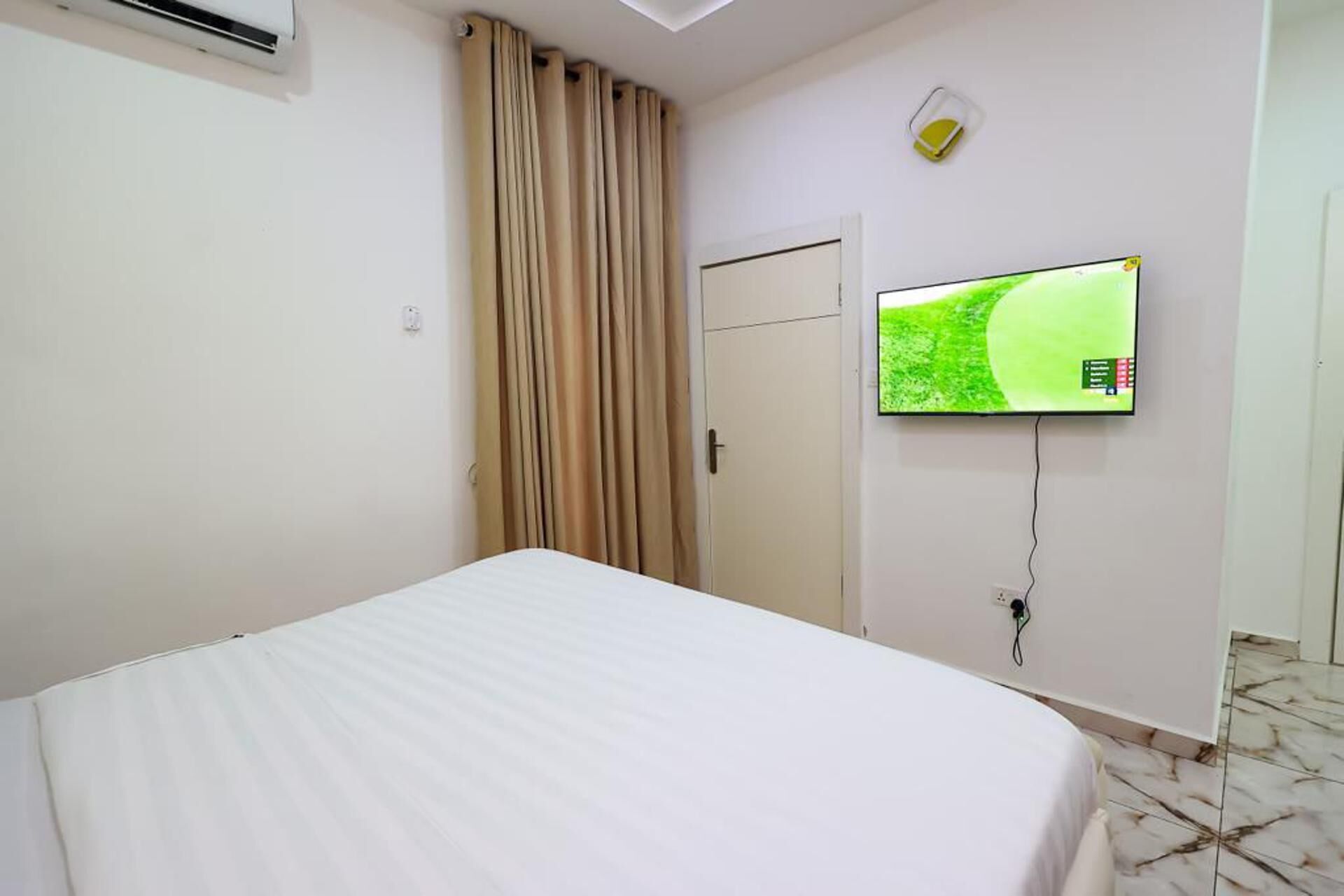 Une chambre d’hôtel avec un lit, un téléviseur fixé au mur, une porte et une fenêtre avec des rideaux.