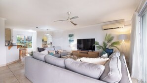 Ein modernes Wohnzimmer mit grauem Sofa, einem Flachbildfernseher, einem Deckenventilator und einem Essbereich mit Tisch und Stühlen.