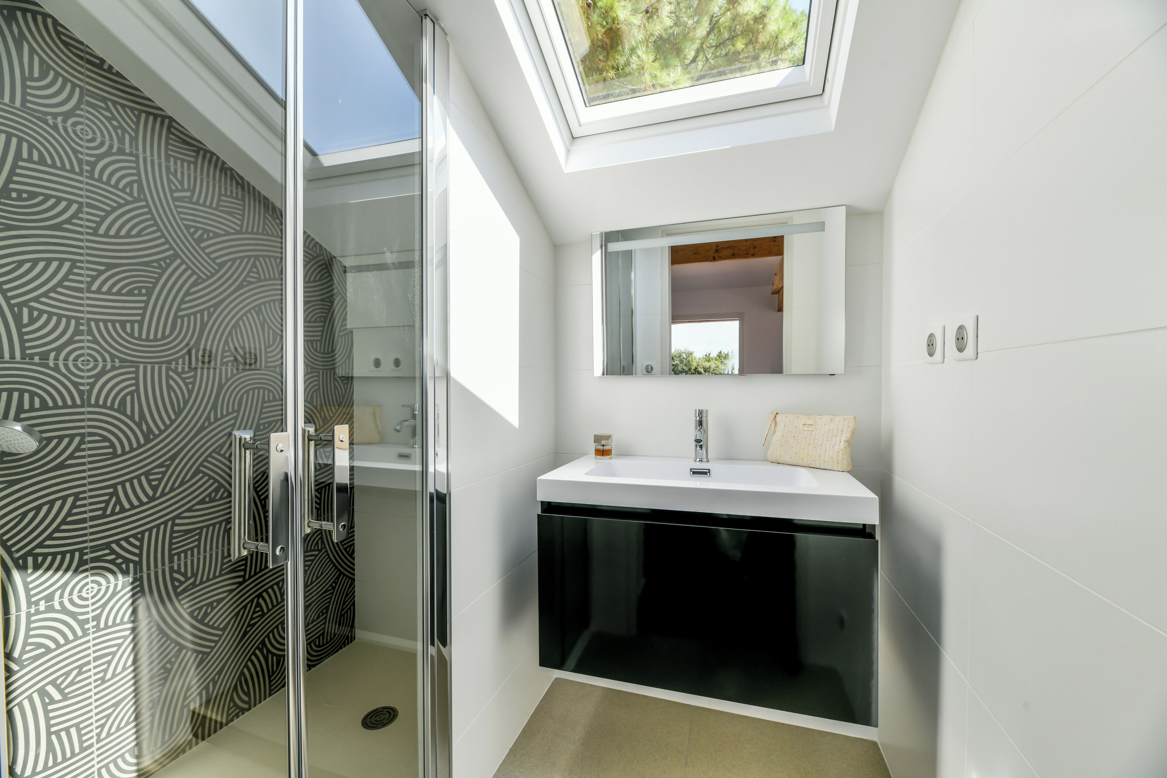 Un baño moderno con tragaluz, cabina de ducha de vidrio y tocador blanco con espejo.