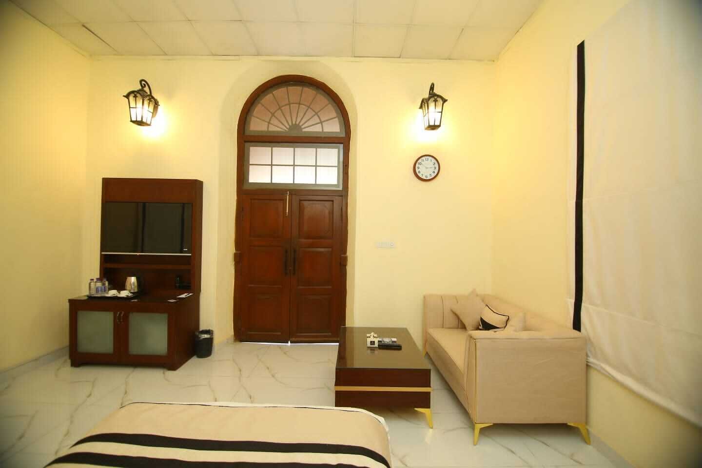 A hotel room with a wooden wardrobe, a television, a sofa, a coffee table, and two wall-mounted lamps.