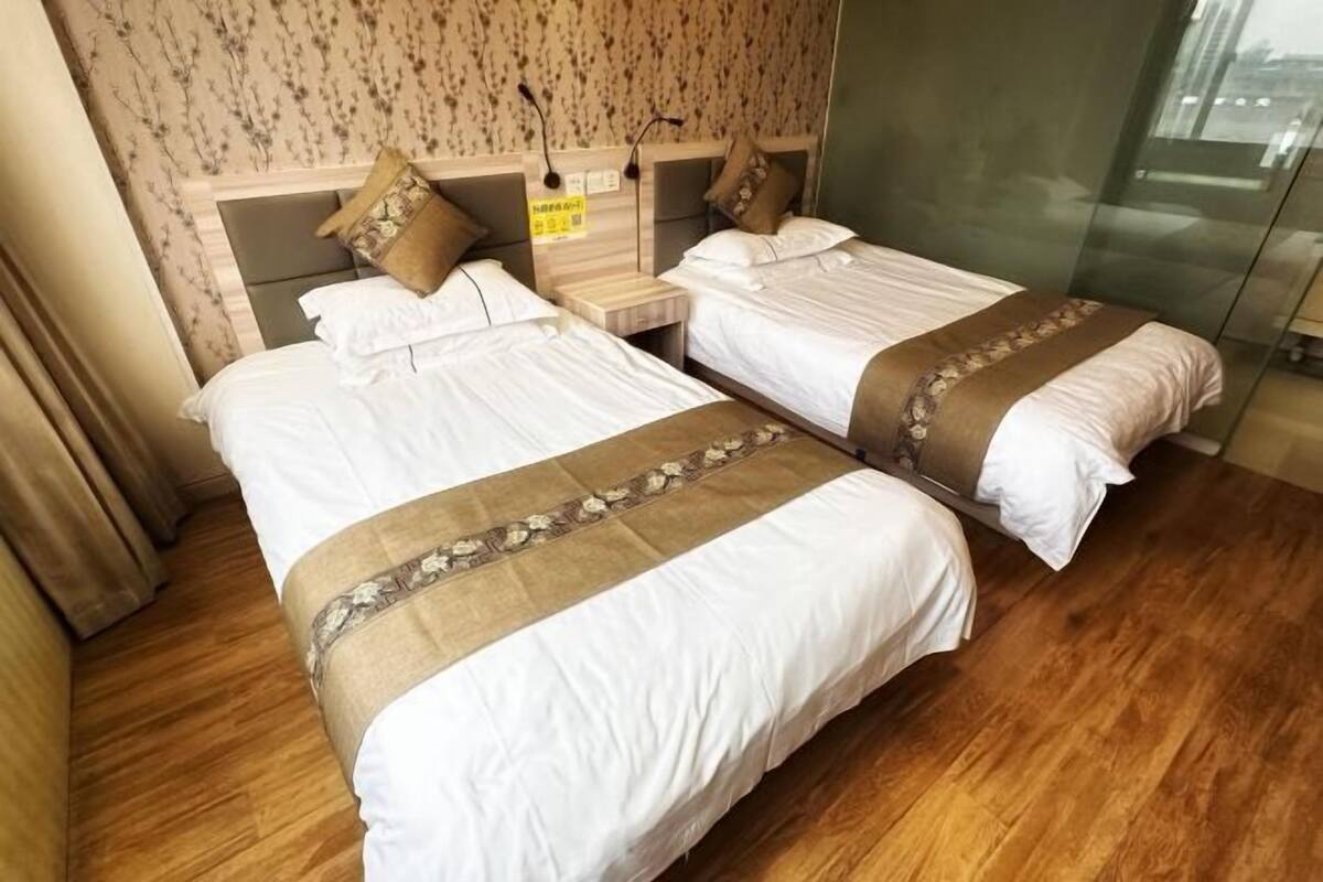 A hotel room with two beds, wooden flooring, floral wallpaper, and a glass partition.