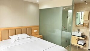 A modern hotel room with a large bed, a glass-enclosed shower, a vanity with a mirror, and a built-in headboard. - 0576 Business Hotel (Taizhou)