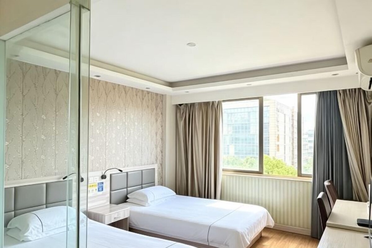 Une chambre d’hôtel avec deux lits, un bureau, une chaise, une douche en verre et une fenêtre avec des rideaux.