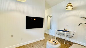 A modern living room with a flat-screen TV mounted on the wall, a round ottoman with a remote, a grey sofa, and a dining area with a table and chairs. - Santorini Cabana in Zephyrhills (Zephyrhills)