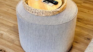 A woven basket on a cylindrical ottoman with a remote control and a bottle. - Santorini Cabana in Zephyrhills (Zephyrhills)