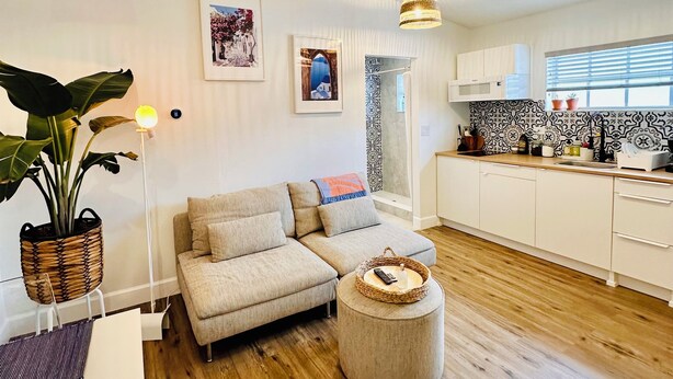 A modern living room with a sofa, ottoman, and a dining table. There is a kitchen area with white cabinets and a backsplash. - Santorini Cabana in Zephyrhills (Zephyrhills)