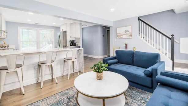 Una sala de estar moderna con un sofá azul, una mesa de centro blanca y una cocina con armarios blancos y una barra.
