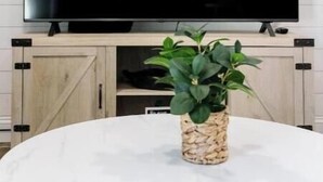 Una sala de estar moderna con un televisor de pantalla plana grande montado en una pared blanca, un mueble de madera debajo y una mesa redonda con una planta en maceta al frente.