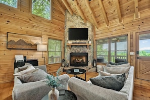 A cozy cabin interior with a stone fireplace, wooden walls, and a flat-screen TV mounted above it.