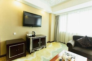 A hotel room with a flat-screen TV mounted on the wall, a dark wooden cabinet with a coffee maker, a sofa with a patterned cushion, and a coffee table with a tray of items.