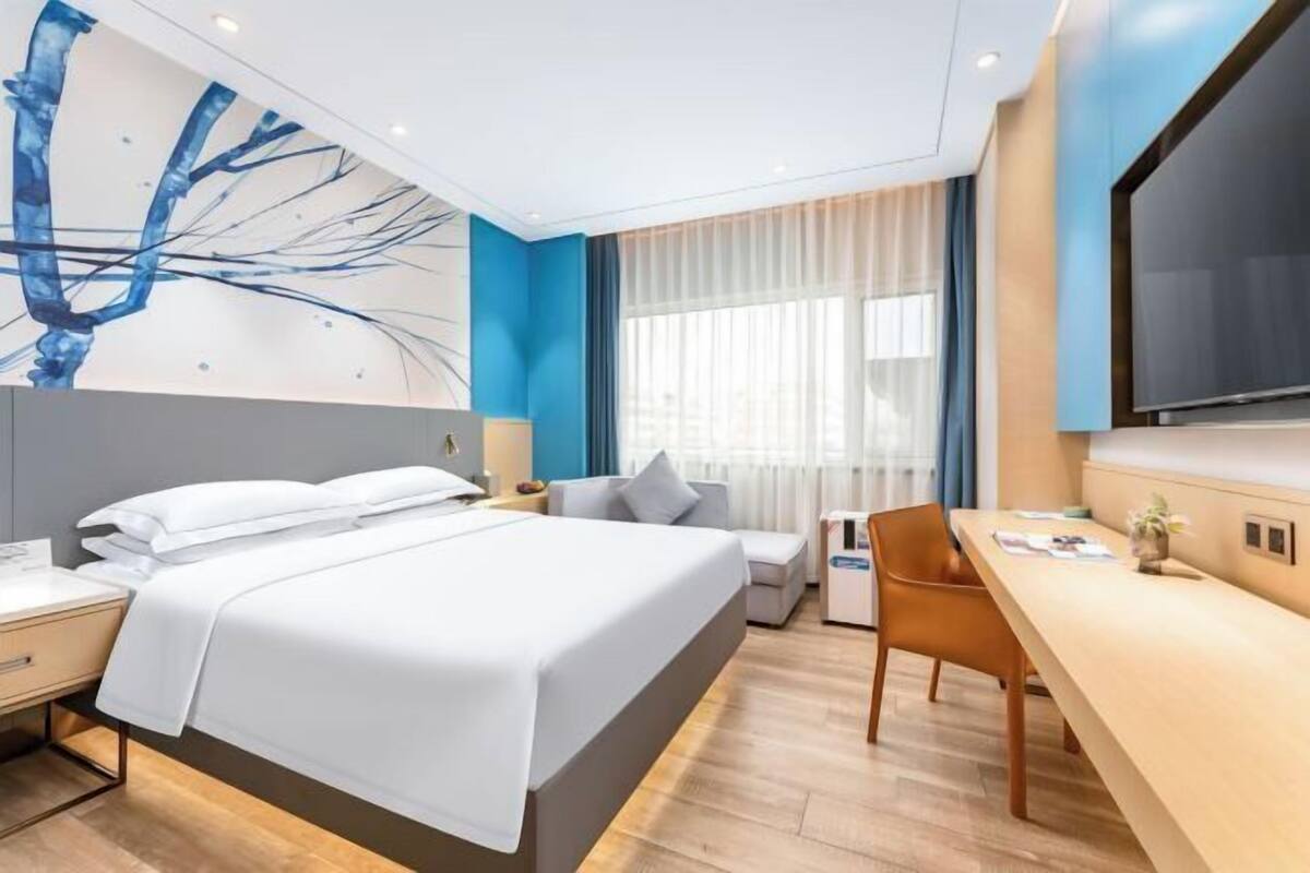 A modern hotel room with a bed, bedside table, chair, desk, and a large TV.