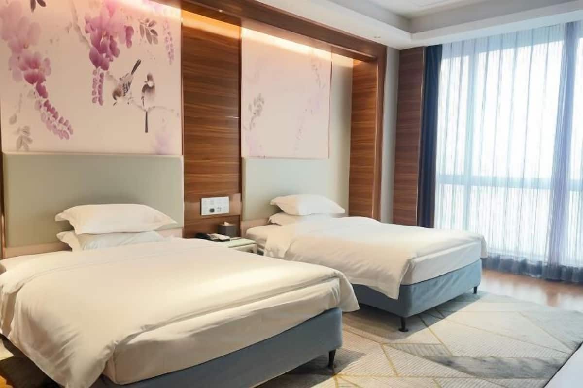 A hotel room with two beds, a large window with sheer curtains, and a wall with floral artwork.