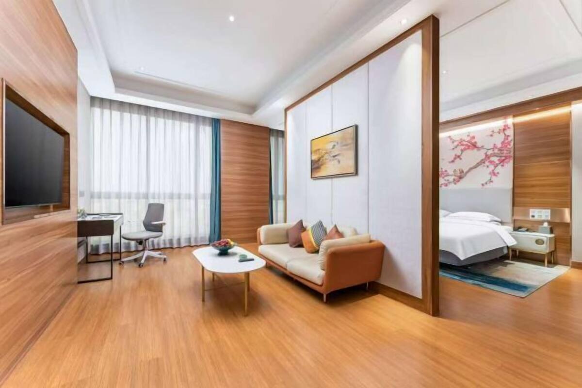 A modern hotel room with a wooden floor, a sofa, a desk with a chair, a bed, and a nightstand.