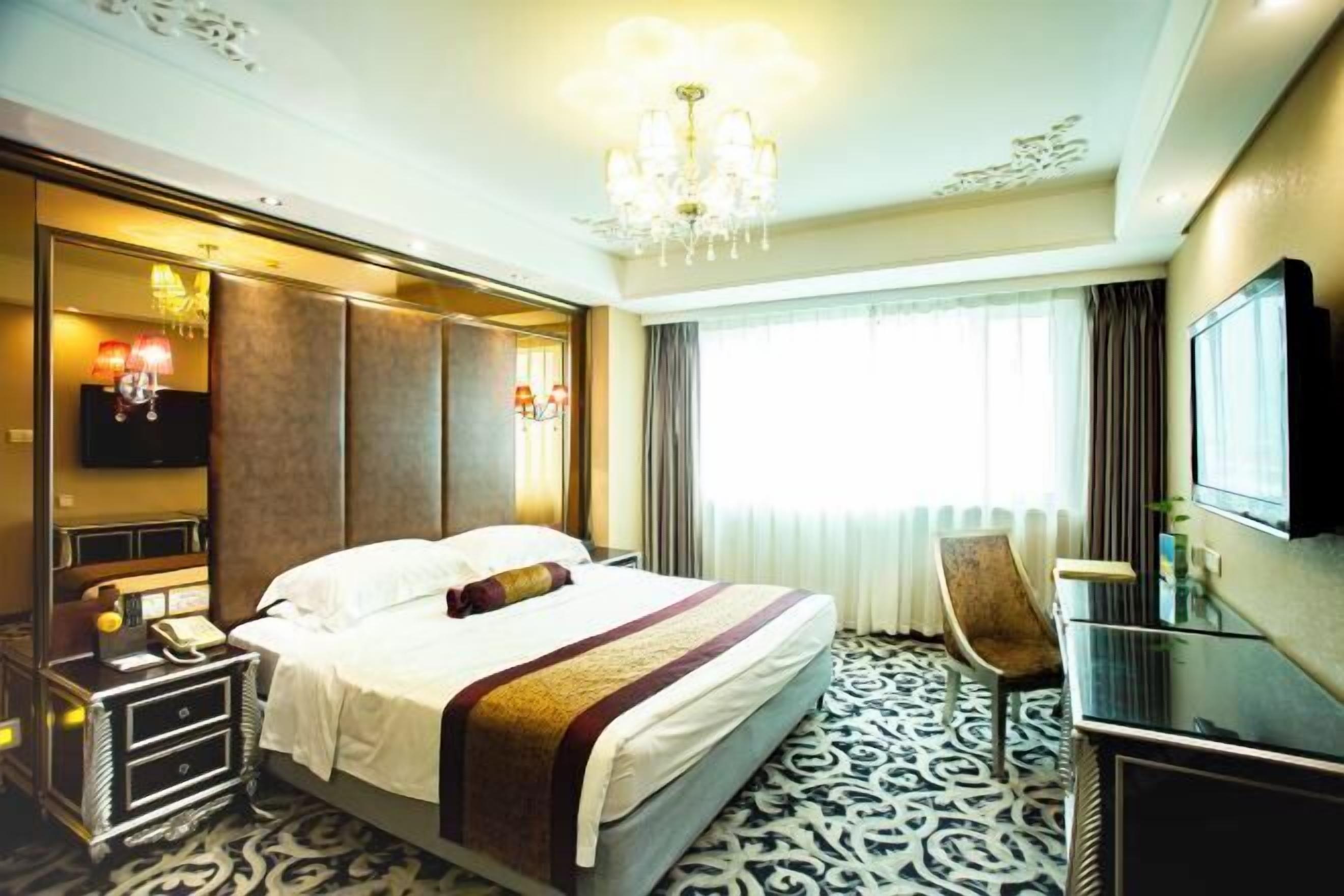 A hotel room with a large bed, a desk, a chair, a TV, and a chandelier.
