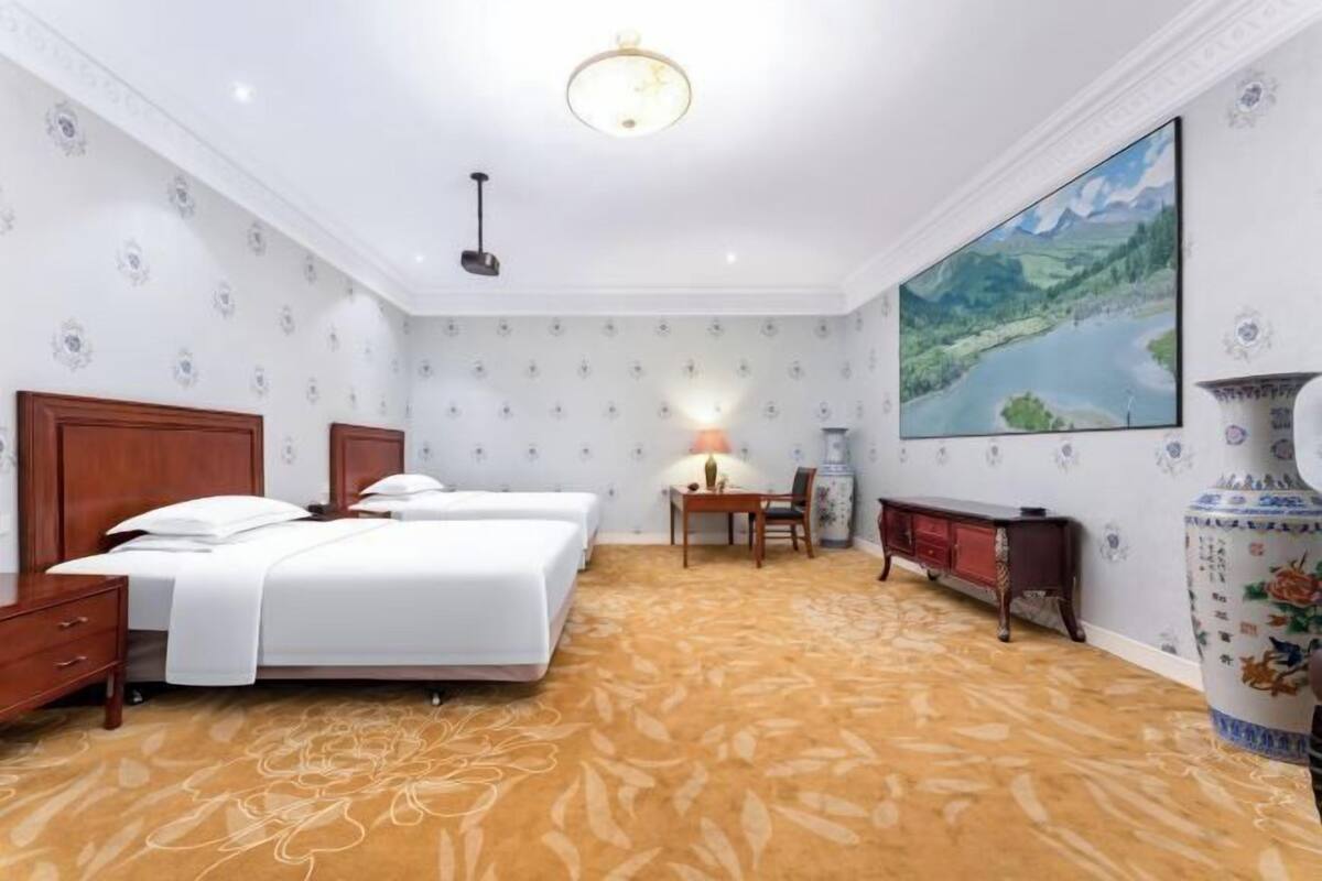 A hotel room with two beds, a desk, a chair, a lamp, and a large painting on the wall.