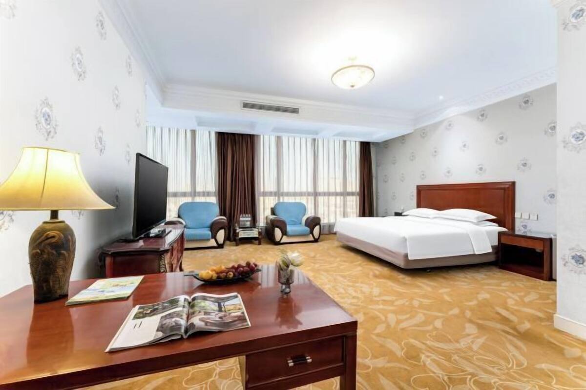 A hotel room with a bed, two armchairs, a desk with a lamp, a television, and a window with curtains.