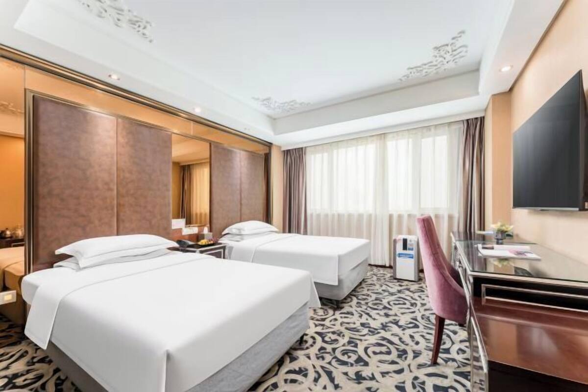 A hotel room with two beds, a desk, a chair, a TV, and a large window with curtains.