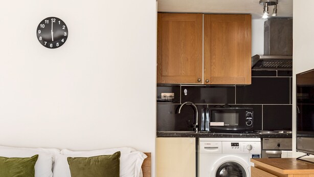 A compact room with a bed, a washing machine, a microwave, and wooden cabinets. - Cosy Studio in Earls Court Flat 17 (London)
