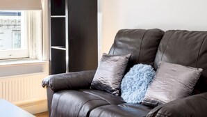 A brown leather sofa with two pillows, a tall black cabinet, and a window with blinds. - Cosy Studio in Earls Court Flat 17 (London)