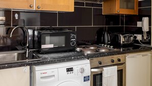 A compact kitchen with wooden cabinets, a black microwave, a Bosch washing machine, and a stove. - Cosy Studio in Earls Court Flat 17 (London)