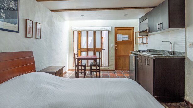Un dormitorio con cama, cabecero de madera, una pequeña mesa con sillas, una puerta de madera y una zona de cocina con armarios y un lavabo.