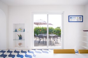 Una habitación con puerta de cristal corredera que da a un patio exterior con mesa y sillas, una planta en maceta y una fotografía en el marco en la pared.