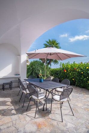 Un patio al aire libre con mesa y sillas, un paraguas grande y vista a un jardín con palmeras y arbustos florecientes.