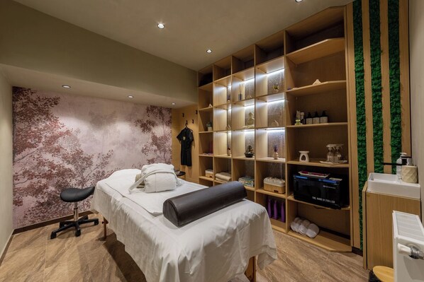 Habitación de hotel con cama, mesa de masaje, estantería de madera grande con varios objetos y pared con un mural de árbol.