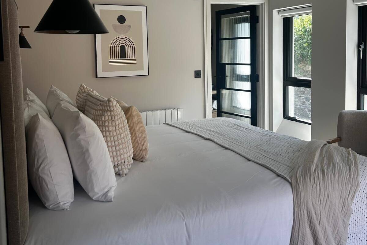 A bedroom with a large bed, a hanging light fixture, a framed picture on the wall, and a window with a view of greenery outside.