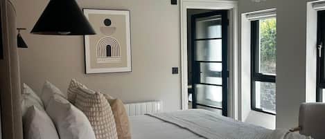 A bedroom with a large bed, a hanging light fixture, a framed picture on the wall, and a window with a view of greenery outside.