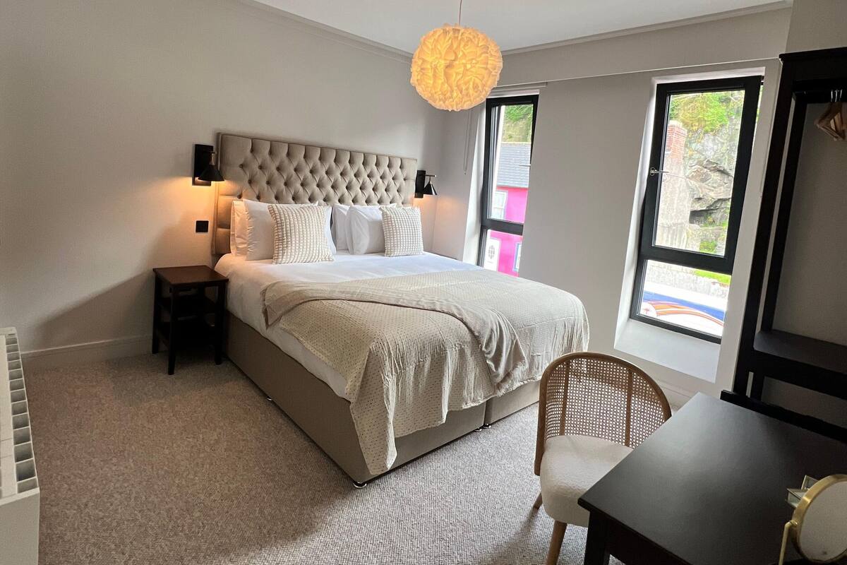 A bedroom with a large bed, a small table, a chair, and a hanging light fixture.