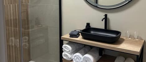 Un baño moderno con cabina de ducha de vidrio, un espejo redondo, un lavamanos negro y toallas ordenadas sobre una repisa de madera.