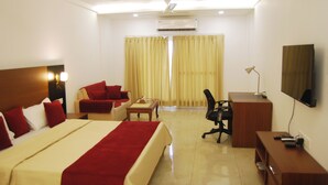 A hotel room with a large bed, a desk, a chair, a TV, and a window with curtains. - Grey Pelican Tourist Resort (Yamunanagar)
