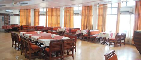 A spacious dining area with multiple tables and chairs, air conditioning units, and large windows with curtains.