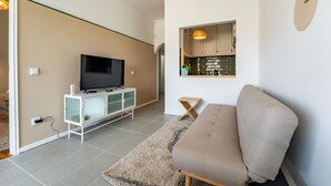 A modern living room with a flat-screen TV on a white cabinet, a beige sofa, and a kitchen area with wooden cabinets and a hanging light fixture.