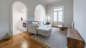 A spacious living area with a sofa, a coffee table, and a wooden bench. There is a kitchen area with a marble countertop and an oven. The room has wooden flooring and white walls. - Nomad s Family - 3bdr City Vibe Porto (Porto)