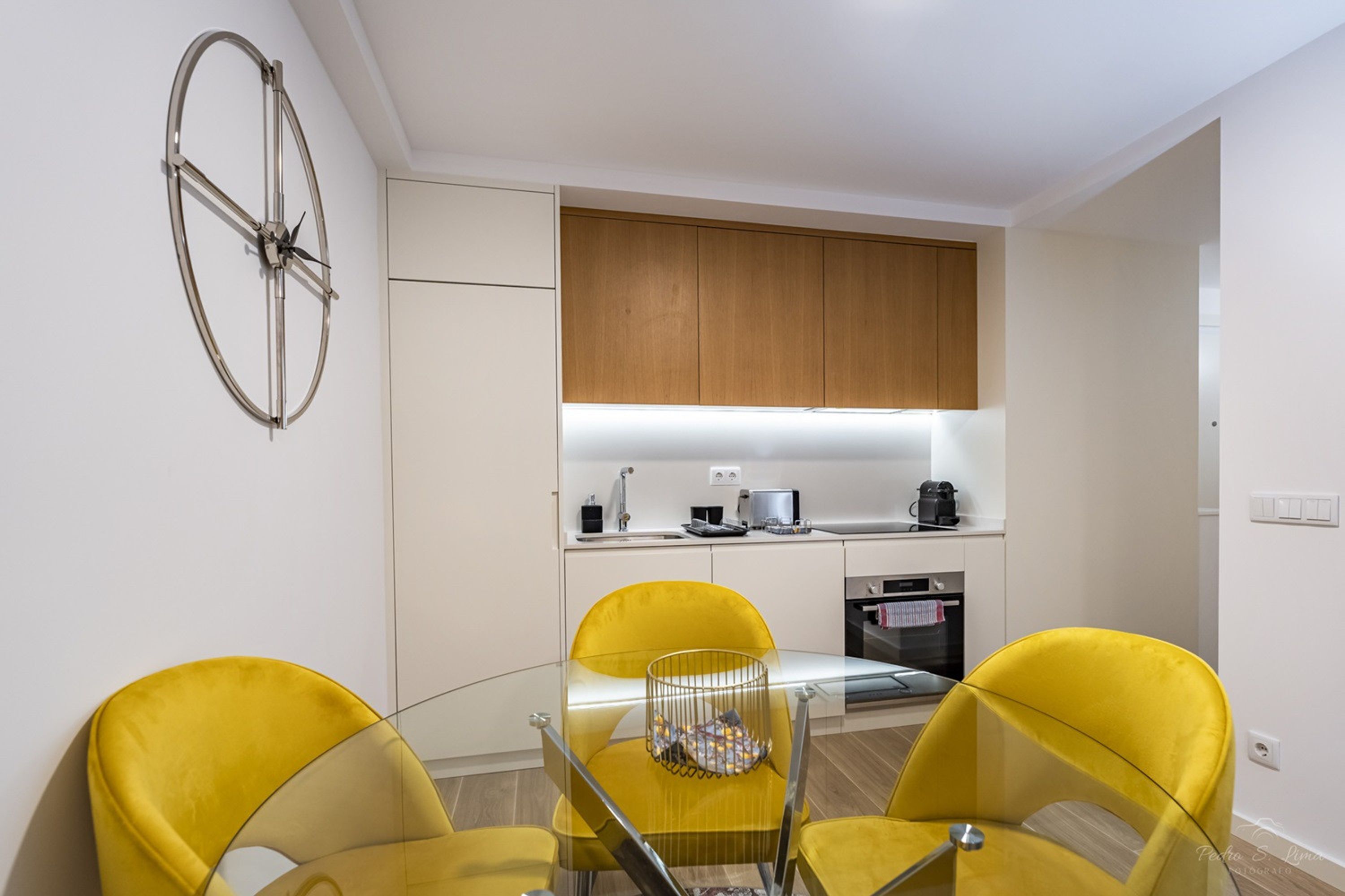 Une cuisine moderne avec des meubles en bois, un four encastré et un petit coin repas comprenant une table en verre et des chaises jaunes.