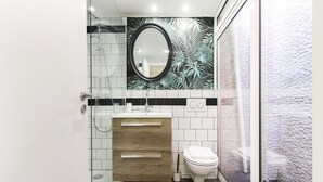 Un baño moderno con ducha envuelta en vidrio, tocador de madera, inodoro blanco y espejo decorativo con detalles florales.