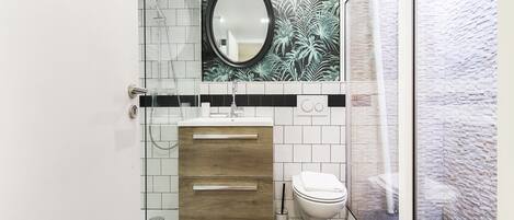 A modern bathroom with a glass-enclosed shower, a wooden vanity, a white toilet, and a decorative mirror with a floral backsplash.