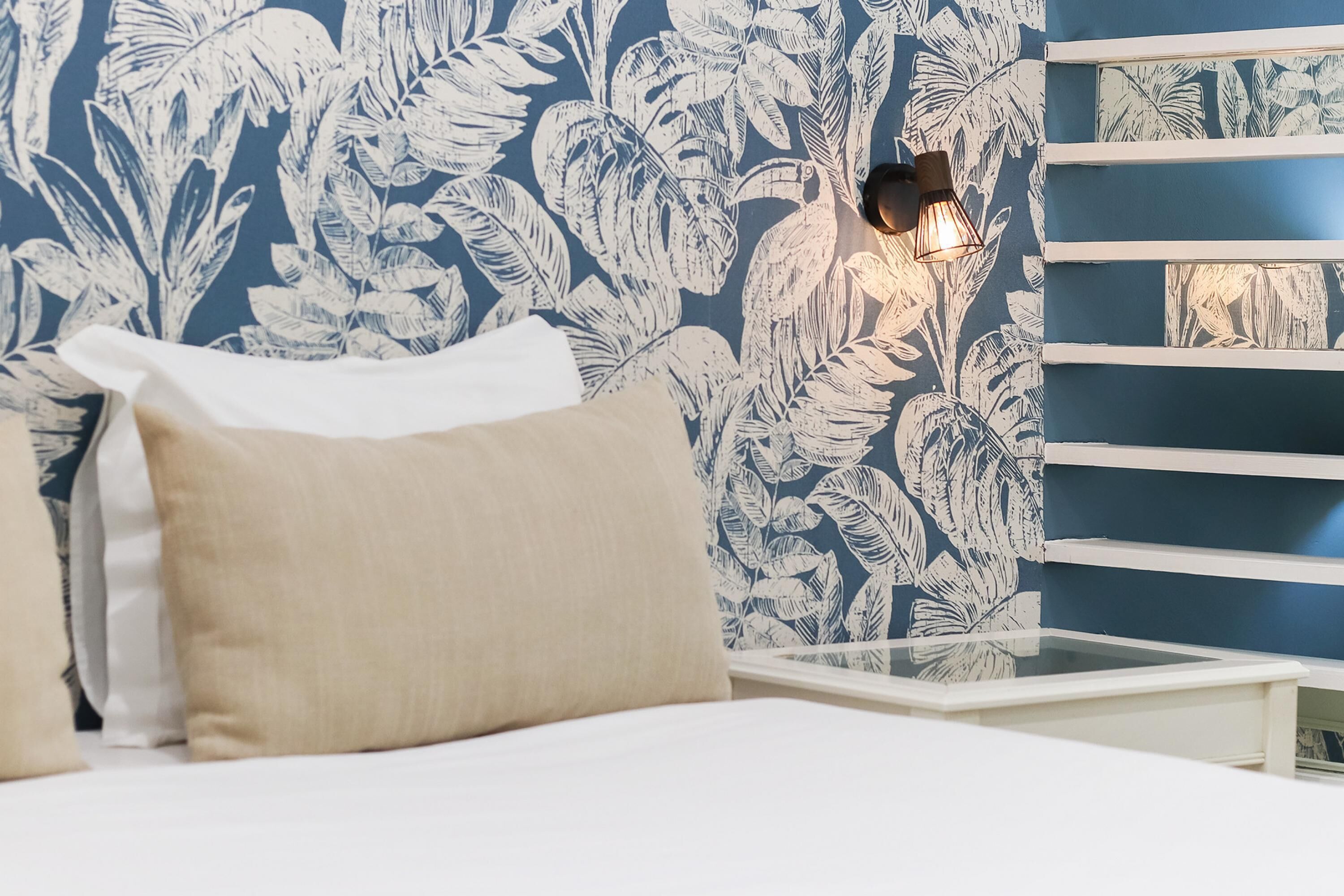 A bedroom with a bed, pillows, a nightstand, a wall-mounted lamp, and a shelf with a blue wall and leaf-pattern wallpaper.
