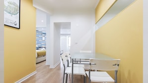 Un interior moderno con comedor que incluye una mesa de vidrio y sillas de metal, una pared de acento amarilla y una cocina visible al fondo.