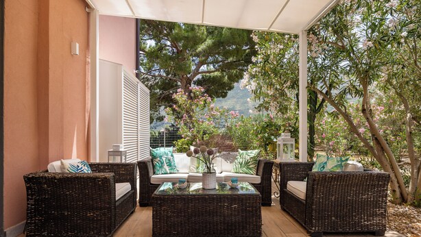 A covered outdoor seating area with wicker furniture and a glass coffee table, overlooking a garden with trees and a mountain view. - Villa Giovanna con Piscina by Wonderful Italy (Castellammare del Golfo)