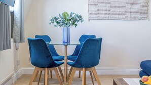 A dining area with a glass table surrounded by four blue chairs, a vase with green foliage, and a woven wall hanging. - Battersea Park 7 Min walk • Balcony • Cafés Below (London)