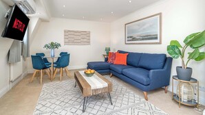 A modern living room with a blue sofa, a dining table with chairs, a coffee table with fruit, a TV on the wall, and a large painting. - Battersea Park 7 Min walk • Balcony • Cafés Below (London)