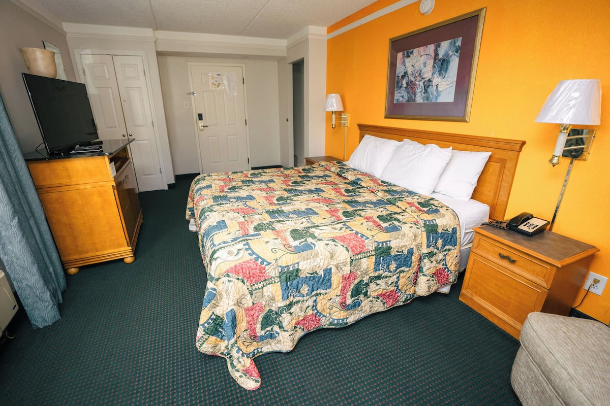 A hotel room with a large bed, a wooden nightstand, a desk with a lamp, a TV on a dresser, and a door leading to another room.