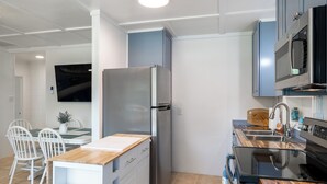 A modern kitchen with a stainless steel refrigerator, a microwave, and a stove. There is a dining area with white chairs and a table. - Bogue Bungalow: Oceanside Duplex, downtown Emerald Isle! (Emerald Isle)