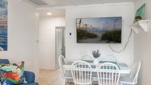 A dining area with a white table and chairs, a painting of a beach scene, and a potted plant on a shelf. - Bogue Bungalow: Oceanside Duplex, downtown Emerald Isle! (Emerald Isle)