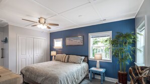 A bedroom with a bed, a ceiling fan, a window with a view, a bedside table, and a chair. - Bogue Bungalow: Oceanside Duplex, downtown Emerald Isle! (Emerald Isle)